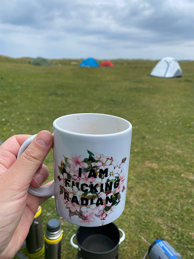 Photograph of Sarah Scott life coach holding a mug of tea at a campsite in the Outer Hebrides. In the background are four small tents on a green grassy field. The sky is cloudy.